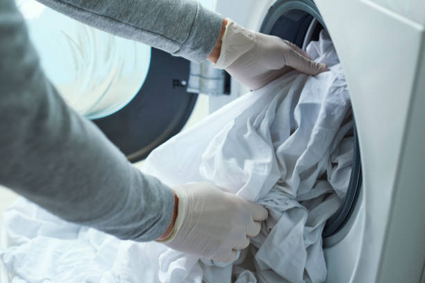 Person wearing gloves placing laundry into a washing machine for sanitizing and disinfecting purposes.